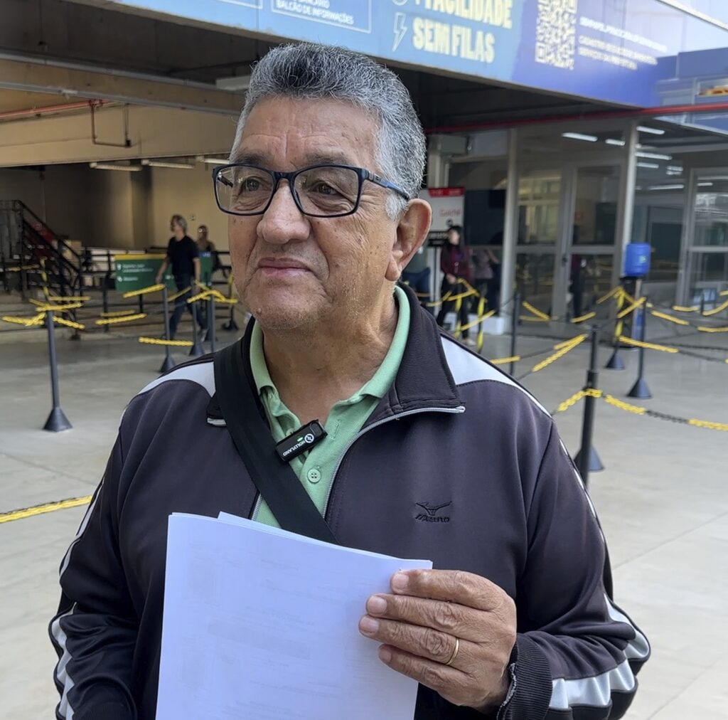 A imagem apresenta um homem de meia-idade, com cabelos grisalhos curtos, usando óculos de armação preta e uma camisa polo verde clara por baixo de uma jaqueta esportiva preta com detalhes brancos nas mangas. Ele segura algumas folhas de papel branco na mão esquerda e possui um pequeno microfone de lapela preso à gola da camisa. O homem está posicionado em primeiro plano, olhando para o lado, com uma expressão serena. Ao fundo, observa-se um ambiente público, possivelmente um centro de atendimento ou repartição governamental, caracterizado por um piso claro, faixas de isolamento amarelas e pretas para organizar filas, e uma estrutura de vidro ao fundo. No alto, uma placa azul exibe a frase "FACILIDADE SEM FILAS" e um código QR, indicando um local voltado para serviços digitais ou atendimento otimizado. Outras pessoas podem ser vistas desfocadas ao fundo, caminhando pelo espaço.