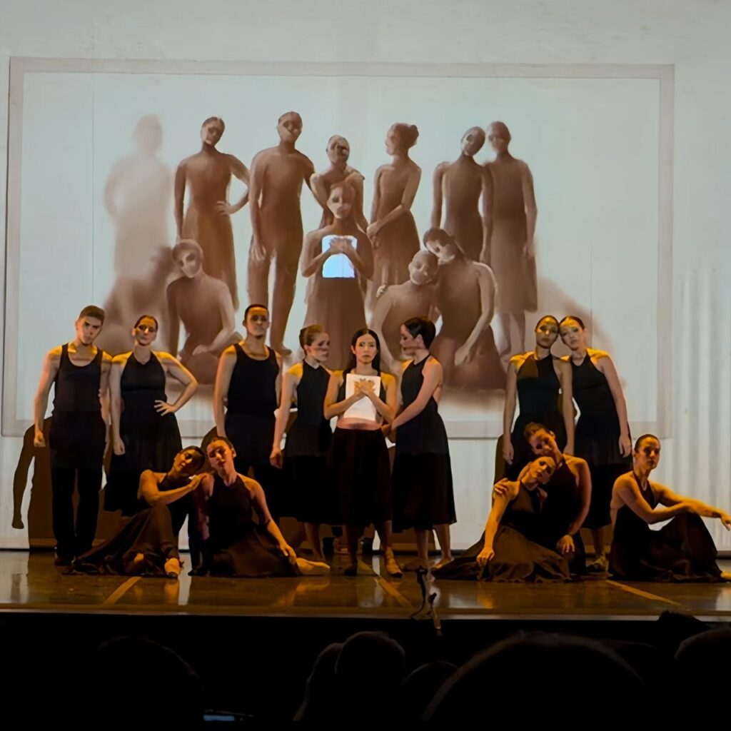 A imagem mostra uma apresentação teatral ou performance de dança em um palco. Ao centro, há um grupo de cerca de 14 pessoas vestindo roupas pretas, com blusas regatas e saias ou calças escuras. Algumas estão em pé e outras sentadas, formando uma composição harmoniosa e equilibrada. Ao fundo, há uma grande tela que projeta uma imagem artística em tons sépia, que parece refletir ou complementar a disposição dos performers no palco. A iluminação é suave e direcionada principalmente para o centro do palco, destacando os artistas. Um dos performers, posicionado no centro, segura um objeto branco que brilha, servindo como ponto focal da cena. O ambiente transmite uma atmosfera artística, séria e contemplativa, com um piso escuro que reflete levemente a luz. A perspectiva da foto é do ponto de vista da plateia, olhando para o palco.