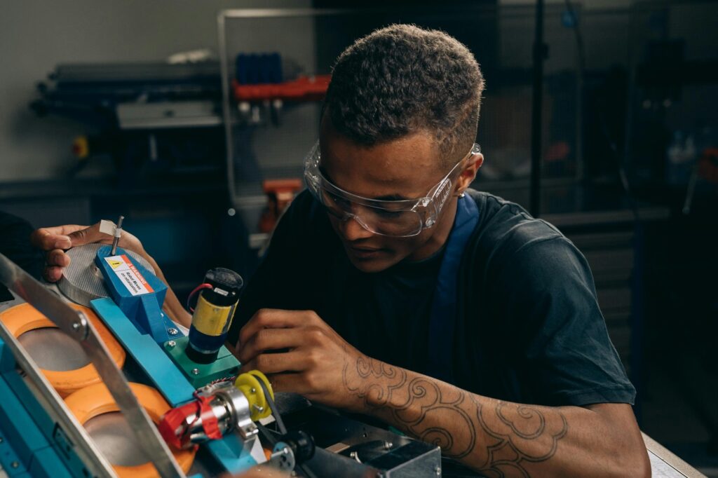 A imagem mostra um jovem concentrado, utilizando óculos de proteção, enquanto trabalha cuidadosamente na montagem ou reparo de um dispositivo mecânico, que parece ser um robô. Ele está em um ambiente que lembra uma oficina ou laboratório, com ferramentas desfocadas ao fundo. O robô possui partes coloridas em tons de azul, laranja e amarelo, e há uma etiqueta de aviso que diz: "WARNING Robot Moves on initialization" (Aviso: o robô se move ao ser iniciado). A iluminação destaca o jovem e o robô, evidenciando o cuidado e a habilidade técnica do trabalho que está sendo realizado.