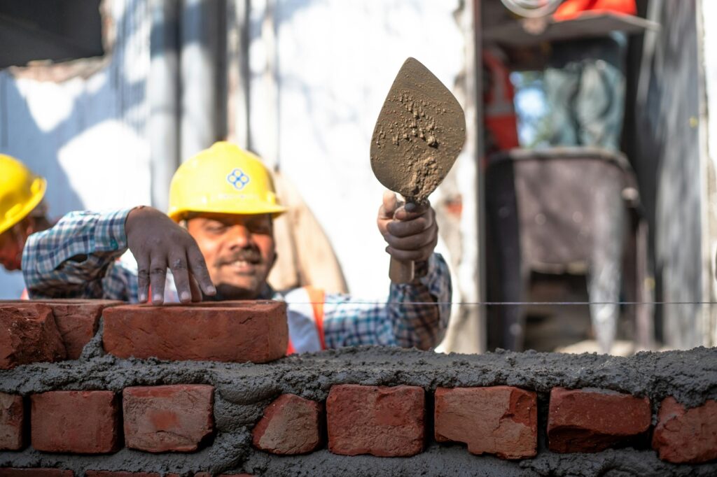 A imagem mostra um trabalhador da construção civil no momento em que está assentando tijolos para levantar uma parede. Ele usa um capacete amarelo de proteção e uma camisa xadrez azul com branco. O homem tem bigode e está concentrado na tarefa, segurando um tijolo vermelho com a mão direita e uma colher de pedreiro com argamassa na mão esquerda. A parede que ele está construindo já tem várias fileiras de tijolos assentados com argamassa cinza. Ao fundo, é possível ver outro trabalhador também usando capacete amarelo, mas ele está desfocado. A cena parece ser ao ar livre, em um canteiro de obras, com iluminação natural suave. A imagem destaca o trabalho manual e a técnica envolvida na construção.