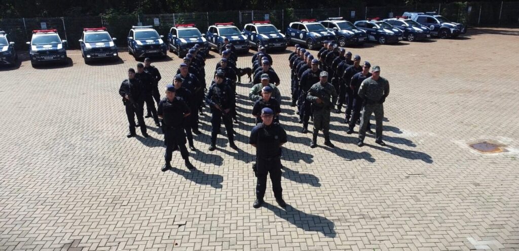 A imagem mostra um grupo de agentes da Guarda Civil Municipal (GCM) perfilados com muita disciplina em um pátio, com várias viaturas policiais estacionadas logo atrás deles. É uma cena que transmite bastante prontidão e organização da equipe! 👮‍♂️🚔 Pelo que dá para ver, eles estão uniformizados e prontos para o serviço, com os veículos devidamente equipados e posicionados em uma formação bem alinhada. Parece ser um dia de sol forte, o que destaca ainda mais o trabalho e a presença deles no local.