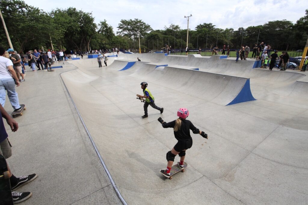 A imagem mostra um parque de skate ao ar livre com uma área de concreto ampla e ondulada, projetada para a prática do skate. No centro da imagem, uma menina com capacete rosa, roupas pretas e equipamentos de proteção está andando de skate em direção ao fundo da foto. À esquerda dela, um menino com um colete refletivo amarelo e capacete preto corre segurando seu skate. Ao fundo, há uma grande estrutura em formato de tigela (bowl) com rampas pintadas de azul, onde várias pessoas estão espalhadas, algumas em pé e outras sentadas, provavelmente observando ou esperando sua vez. O ambiente está cercado por árvores altas, indicando que o parque está em uma área verde. O céu está nublado, o que deixa a iluminação suave e sem sombras fortes. O cenário transmite um momento de lazer e atividade física em grupo, com crianças e adultos aproveitando o espaço.