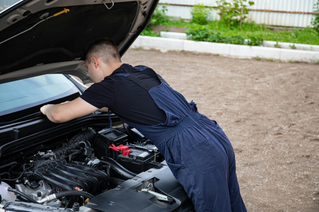 A imagem mostra um homem vestindo um macacão azul sobre uma camiseta preta, inclinado sobre o motor de um carro com o capô aberto. Ele está de costas para a câmera e parece estar trabalhando ou inspecionando o motor. O ambiente é externo, com chão de terra ou cascalho, uma mureta branca ao fundo e vegetação ao longe. A cena transmite a ideia de manutenção ou reparo automotivo, com foco na interação do homem com o veículo.