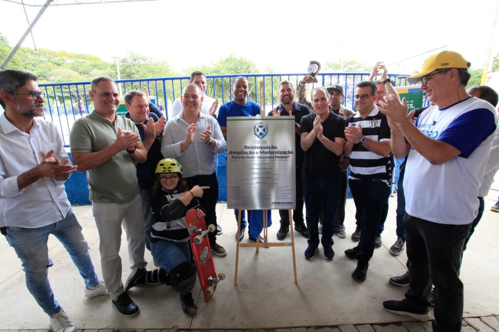 A imagem mostra a inauguração ou reabertura de uma pista de skate chamada "Alexandre Peverari", localizada em Piracicaba. No centro da foto, há uma placa comemorativa sobre um cavalete de madeira, com os seguintes dizeres: "Readequação, Ampliação e Modernização Pista de Skate 'Alexandre Peverari'". A placa também menciona o prefeito municipal Luciano Almeida, a vice-prefeita Érica Ferrarin e o secretário municipal de esportes, lazer e atividades motoras Cléber Canteiro, datada de maio de 2024. Em frente à placa, uma criança ou adolescente usando capacete amarelo, joelheiras e segurando um skate vermelho está agachada, apontando para a placa. Ao redor dela, há um grupo de homens adultos, possivelmente autoridades ou organizadores, que estão aplaudindo e sorrindo. Eles estão vestidos de forma casual, com camisas polo, camisas sociais e jeans. O ambiente parece ser externo, com uma cerca azul ao fundo e iluminação natural, indicando que o evento ocorre durante o dia. A atmosfera é de celebração e reconhecimento do espaço renovado para a prática do skate.
