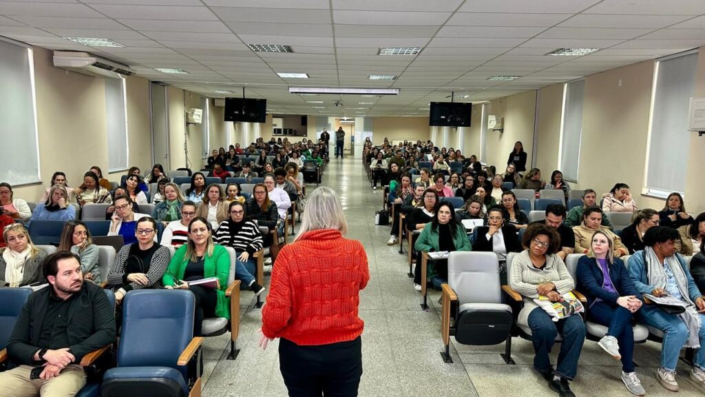 A imagem mostra um auditório grande e bem iluminado, com muitas pessoas sentadas em cadeiras azuis acolchoadas dispostas em fileiras que se estendem ao longo da sala. A maioria das pessoas na plateia são mulheres, vestidas de forma casual e atentas ao que está acontecendo à frente. Em primeiro plano, de costas para a câmera, está uma pessoa de cabelo loiro usando um suéter laranja, aparentemente a palestrante ou facilitadora do evento. A sala tem paredes claras e um teto com painéis brancos, além de duas telas de televisão montadas nas paredes próximas à frente. A atmosfera parece ser profissional, sugerindo que pode ser uma palestra, workshop ou reunião formal.