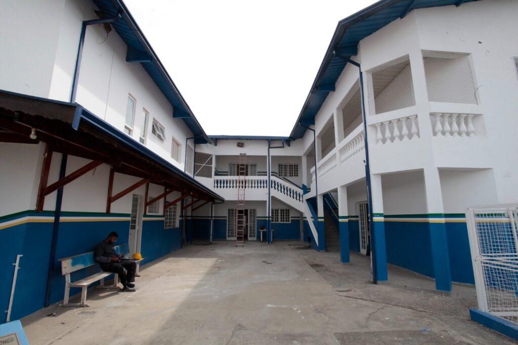 A imagem mostra o pátio interno de um prédio de dois andares, possivelmente uma escola ou um edifício público. O prédio é pintado em branco com detalhes em azul e uma faixa amarela e branca na parte inferior da parede. O telhado é azul e há uma escada de metal vermelha encostada na parede central do prédio ao fundo. No pátio, há um banco de madeira desgastado onde um homem está sentado, usando roupas escuras e um capacete amarelo apoiado ao lado. À direita, há uma escada que leva ao segundo andar, onde há uma varanda com balaústres brancos. No canto inferior direito, há um portão de metal branco, parcialmente aberto. O chão do pátio é de concreto, e a iluminação indica que o dia está nublado, com luz difusa e sem sombras fortes. A atmosfera geral é tranquila e o local parece estar em manutenção ou algum tipo de reforma, sugerido pela presença da escada e do capacete.