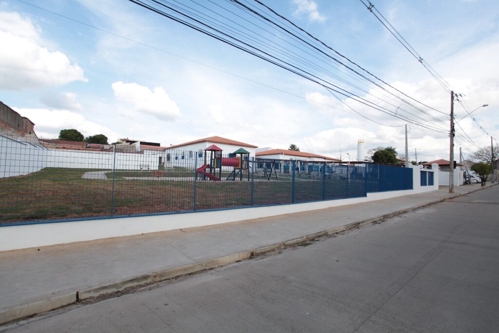 A imagem mostra a fachada de uma escola ou creche, com um muro baixo pintado de azul que cerca o pátio externo. No pátio, há um pequeno playground colorido com escorregador e outros brinquedos para crianças. O prédio é de um andar, com paredes brancas e um telhado de telhas vermelhas, típico de construções em regiões tropicais. O céu está parcialmente nublado, com bastante luz natural iluminando a cena. No primeiro plano, há uma calçada de concreto e vários fios de energia elétrica cruzando o céu. Ao fundo, é possível ver uma torre de água alta. A imagem transmite uma sensação de um ambiente escolar tranquilo e bem cuidado.