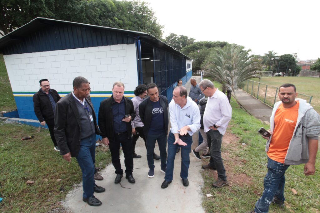A imagem mostra um grupo de homens em pé ao lado de um prédio branco com a parte inferior pintada de azul, separado por uma faixa verde e amarela. Eles parecem estar em uma conversa ou reunião, com um homem no centro fazendo um gesto com as mãos, possivelmente explicando algo. Um dos homens está usando uma camiseta laranja com a palavra "estrela" escrita na frente. O local parece ser uma área externa, com um campo de grama e uma trave de futebol ao fundo, cercado por uma cerca. A atmosfera é de um encontro informal ou uma visita técnica em um ambiente que pode ser escolar ou esportivo.