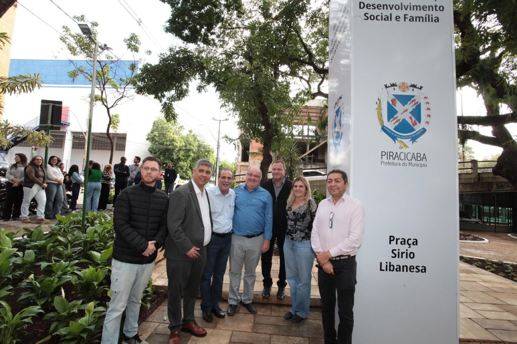 A imagem mostra um grupo de oito pessoas posando para uma foto ao ar livre, em frente a um totem informativo na Praça Sirio Libanesa, em Piracicaba. * O grupo principal, composto por seis homens e uma mulher, está posicionado à direita, ao lado de um totem branco vertical que exibe o brasão da cidade de Piracicaba e o nome do local. * As pessoas vestem roupas casuais e sociais, como camisas, calças jeans, blazers e jaquetas. Elas estão sorrindo ou olhando para a câmera. * À esquerda, um pouco mais distantes, há outras pessoas caminhando ou conversando em uma área arborizada com canteiros de plantas. * O cenário ao fundo revela um ambiente urbano, com árvores frondosas, um edifício em construção e postes de iluminação, sob um céu claro e ensolarado.