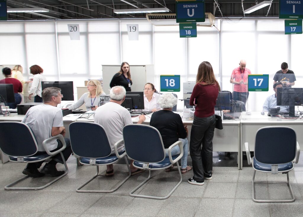 A imagem mostra um ambiente de atendimento ao público, possivelmente uma repartição pública ou central de serviços, com várias mesas dispostas lado a lado. * Os atendentes estão sentados atrás das mesas, interagindo com os clientes. Há divisórias de acrílico transparente entre os atendentes e o público. * Os postos de atendimento são identificados por números suspensos, como 17, 18, 19 e 20. Acima das mesas, placas indicam "SENHAS U" (Unimed) e "SENHAS I" (SEMAE). * Vários cidadãos estão sentados em cadeiras azuis de metal, aguardando ou sendo atendidos. Uma mulher de blusa bordô está em pé, conversando com a atendente do posto 18. * O ambiente é iluminado por luzes fluorescentes no teto, o piso é de granilite cinza e há computadores e outros equipamentos de escritório sobre as mesas. O clima parece ser de um dia de trabalho comum em um setor de atendimento administrativo.