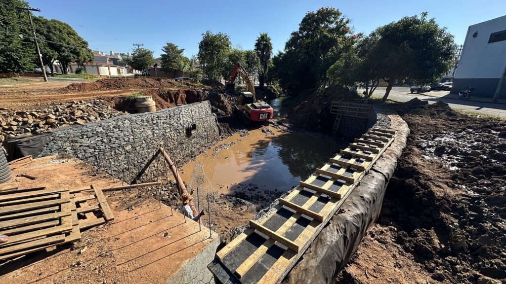 A imagem mostra um canteiro de obras em um dia claro e ensolarado. No centro, há um canal parcialmente cheio de água barrenta, cercado por paredes reforçadas com gabiões — estruturas de malha metálica preenchidas com pedras. Um trator escavador amarelo da marca CAT está posicionado dentro do canal, aparentemente trabalhando na área mais distante da água. Na frente da imagem, há uma estrutura de madeira e painéis pretos que parecem ser uma fôrma para concreto, apoiada sobre uma lona preta no solo. Ao lado esquerdo, há uma pilha de pedras e terra solta. Ao fundo, é possível ver algumas árvores, um trecho de estrada com carros estacionados e um edifício branco. A cena transmite a ideia de uma obra em andamento, possivelmente para reforço ou construção de um sistema de drenagem ou canalização.