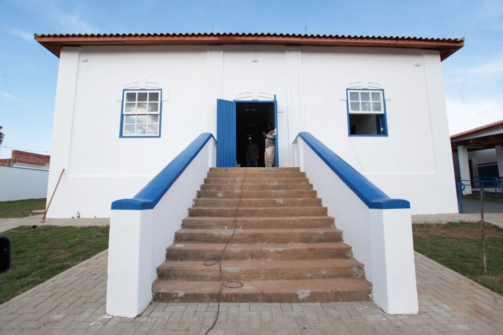 A imagem mostra uma construção de estilo colonial ou tradicional, pintada de branco com detalhes em azul, como as molduras das janelas e a porta central. A fachada é simétrica, com duas janelas de vidro com moldura azul, uma de cada lado da porta aberta, também azul. Para acessar a entrada, há uma escadaria ampla de concreto com corrimões baixos pintados de branco e azul. No interior da entrada, que está em sombra, aparecem duas pessoas, uma delas usando chapéu. O telhado da construção é de telhas vermelhas e o céu está claro e azul, indicando um dia ensolarado. Ao redor da construção, há áreas com grama e um caminho pavimentado com blocos de concreto. Um cabo preto está estendido desde a porta até o chão. A aparência geral do prédio é limpa e bem conservada, sugerindo que pode ser um edifício público, histórico ou religioso que passou por uma restauração recente.
