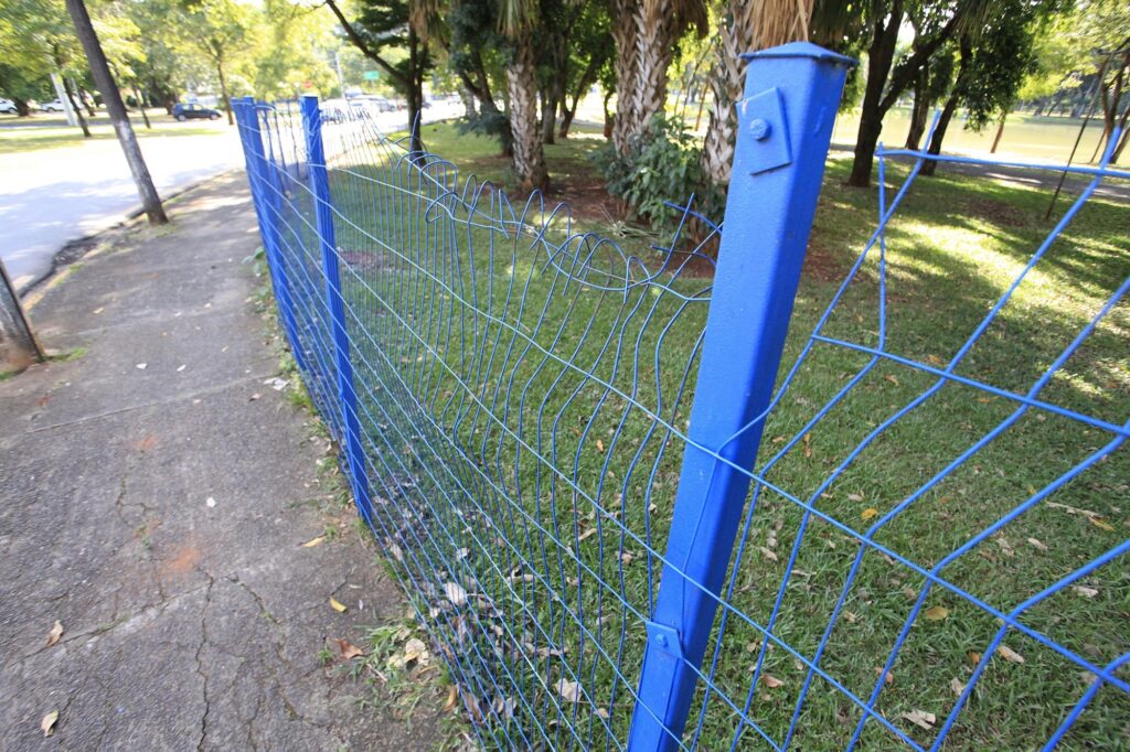 A imagem mostra uma cerca de metal pintada de azul vibrante, com uma malha de arame, que se estende ao longo de uma calçada de concreto. A cerca apresenta sinais de desgaste, especialmente na parte superior, onde os fios estão dobrados e danificados. A perspectiva da foto é em ângulo baixo, destacando a extensão da cerca que se prolonga em direção ao fundo da imagem. À esquerda da cerca, há uma calçada de concreto com rachaduras visíveis, e ao fundo, é possível ver uma rua com um carro estacionado. À direita da cerca, há uma área gramada com algumas palmeiras e outras plantas, sugerindo que o local pode ser um parque ou uma área pública ao ar livre. A iluminação é forte e ensolarada, indicando que a foto foi tirada durante o dia, com sombras bem definidas no chão.