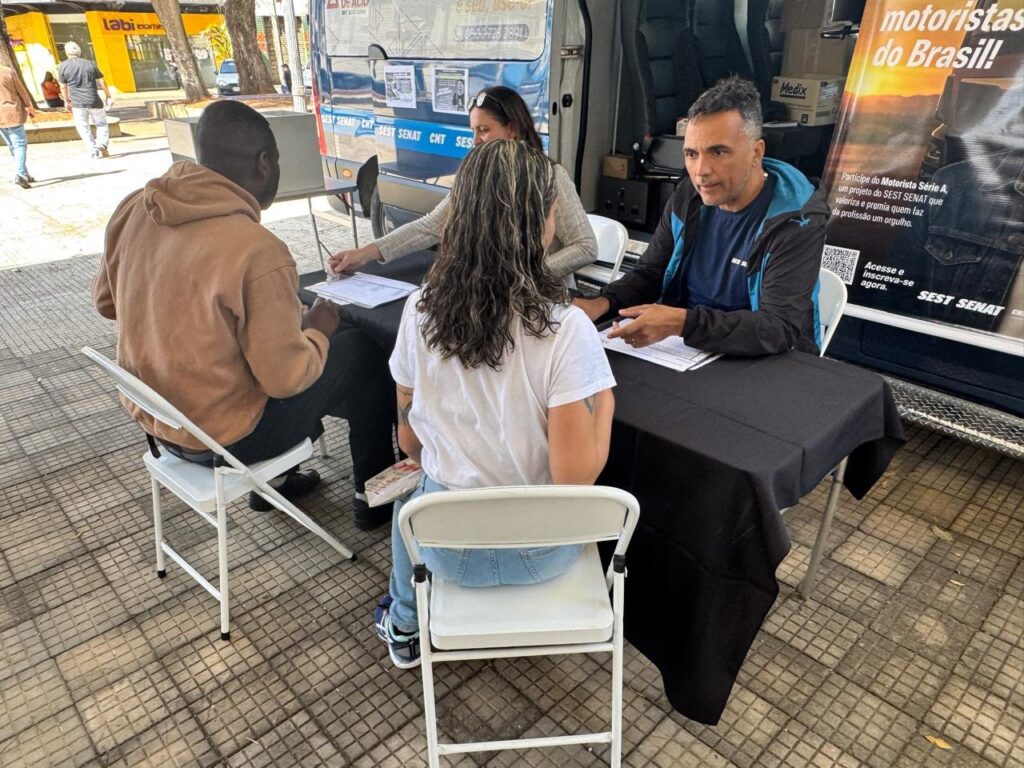 A imagem mostra uma cena ao ar livre, provavelmente em uma calçada ou praça, onde está montada uma mesa para atendimento ou inscrição em um evento ou programa. Na mesa, coberta por uma toalha preta, estão sentadas três pessoas: um homem de moletom bege, uma mulher com cabelo loiro e uma camiseta branca, e um homem com uma jaqueta azul e preta, que parece ser um atendente ou representante do evento. Eles estão analisando documentos juntos. Ao fundo, há uma van azul com a marca "SEST SENAT" e um banner grande com informações sobre o programa "Motorista Série A", que é um projeto do SEST SENAT voltado para motoristas do Brasil, com o objetivo de valorizar e premiar os profissionais da categoria. O banner convida os motoristas a participarem e se inscreverem no projeto. Também é possível ver um prédio amarelo com a placa "Labi exames" e uma pessoa caminhando ao fundo. A cena transmite um ambiente de atendimento e orientação para motoristas interessados no programa.