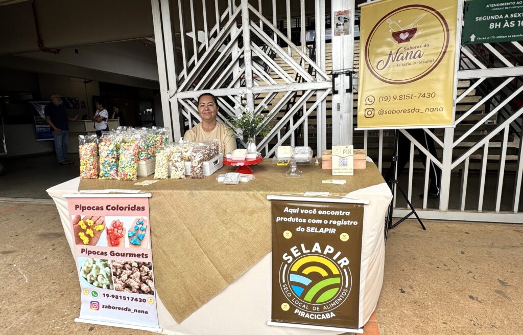 A imagem mostra uma barraca de venda de pipocas artesanais em um ambiente externo, possivelmente uma feira ou mercado local. No centro, há uma mulher sorridente, vestindo uma camiseta marrom clara, atrás de uma mesa coberta com toalhas bege e branca. Sobre a mesa, há diversos sacos plásticos transparentes contendo pipocas coloridas e pipocas gourmet, além de alguns potes e embalagens com outros produtos. À direita da mulher, há uma faixa amarela com o logo "Sabores da Nana Confeitaria Artesanal", informações de contato (telefone e Instagram) e detalhes sobre o horário de funcionamento (segunda a sexta, das 8h às 16h). Na frente da mesa, há dois banners, um rosa e outro marrom, que promovem os produtos e exibem o selo local de alimentos de Piracicaba (SELAPIR), indicando que os produtos são registrados e de origem local. O ambiente parece ser ao ar livre, com chão de concreto e uma estrutura metálica branca ao fundo. A barraca é organizada e transmite uma sensação de cuidado e artesanato nos produtos oferecidos.