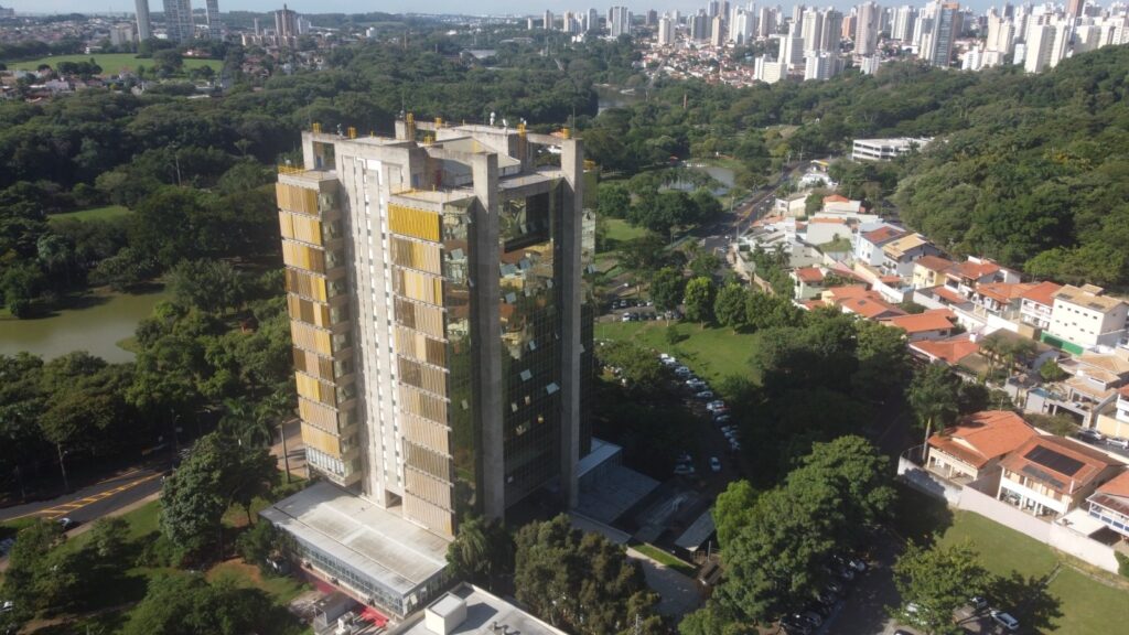 Esta imagem apresenta uma vista aérea, em ângulo zenital, de um edifício comercial moderno de concreto e vidro, localizado em uma área urbana cercada por vegetação densa. * O prédio principal, em destaque, possui uma fachada com estrutura de concreto aparente e painéis verticais amarelos, contrastando com as faces espelhadas que refletem o céu e o entorno. * Ao redor da construção, observa-se um parque arborizado com um lago à esquerda, além de um estacionamento contornando a estrutura. * À direita, estende-se um bairro residencial com casas de telhados cerâmicos tradicionais. * No horizonte, ao fundo, avista-se o centro de uma cidade com diversos edifícios altos, indicando uma área densamente urbanizada. A iluminação sugere um dia claro e ensolarado.