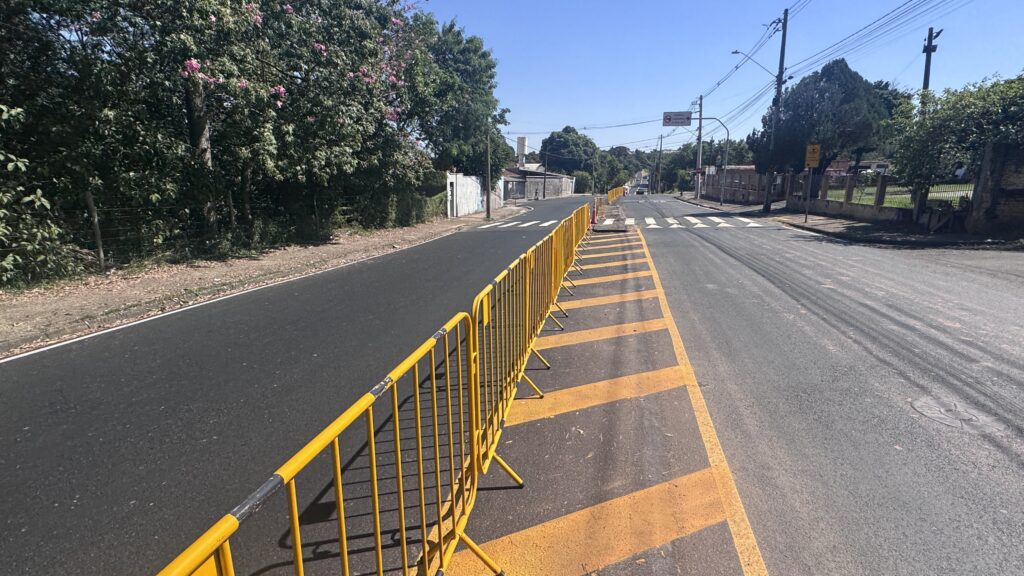 A imagem mostra uma rua em um dia ensolarado, com um céu azul claro e árvores verdes ao longo do lado esquerdo da via. Ao longo da rua, há várias barreiras amarelas de metal que formam uma espécie de proteção ou isolamento, provavelmente para controle de tráfego ou para sinalizar uma obra em andamento. O asfalto da rua é escuro e parece estar em boas condições, com algumas faixas amarelas pintadas no chão que indicam limites ou orientação para os veículos. Do lado direito da imagem, há postes de luz e algumas construções residenciais ou comerciais ao fundo. Também é possível ver uma placa de sinalização com o texto em português "PROIBIDO CAMINHAR SOBRE O MURO", indicando que não é permitido caminhar sobre um muro próximo, provavelmente por questões de segurança. A perspectiva da foto é baixa, olhando ao longo da rua, o que dá uma sensação de profundidade e mostra o alinhamento das barreiras amarelas que acompanham o caminho da via até o horizonte. A iluminação é forte, com sombras nítidas formadas pelas barreiras no chão. No geral, a imagem transmite uma cena urbana tranquila, com elementos que indicam precauções para pedestres e veículos em uma área possivelmente em manutenção ou com restrições de circulação.
