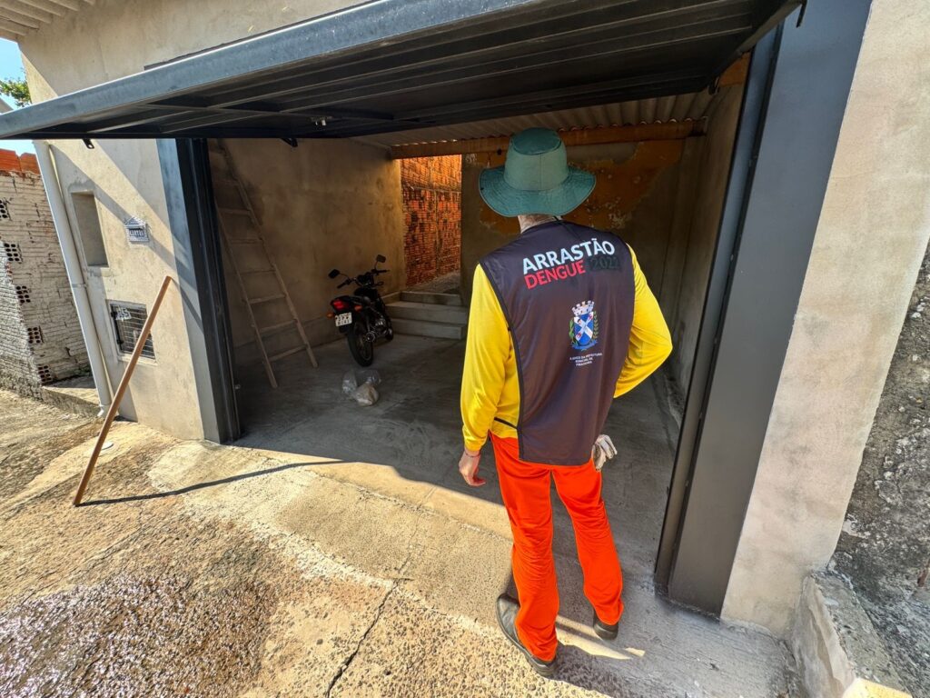 A imagem mostra uma pessoa vista por trás, usando um chapéu verde de aba larga, uma camisa amarela de manga longa, calças laranja e um colete preto com a inscrição "ARRASTÃO DENGUE 2022" e o brasão da Prefeitura Municipal de Piracicaba. A pessoa está na entrada de uma garagem, aparentemente realizando um trabalho de combate à dengue. Dentro da garagem, é possível ver uma motocicleta estacionada e uma escada de madeira encostada na parede. O ambiente parece ser residencial, com piso de concreto e iluminação natural. A imagem transmite a ideia de uma ação pública de saúde voltada para o controle do mosquito transmissor da dengue.
