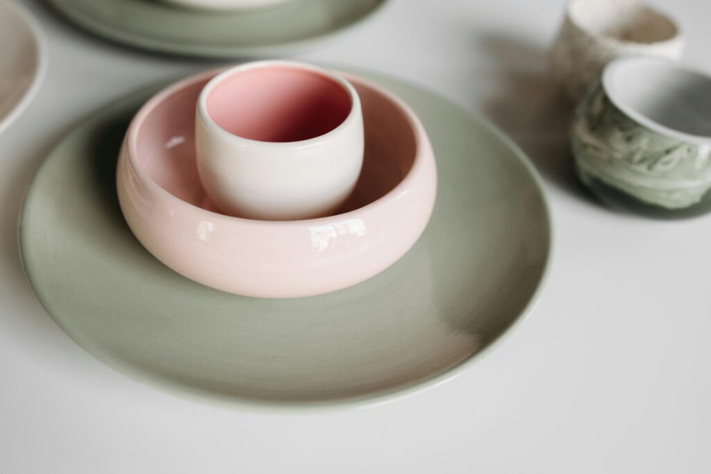 A imagem mostra uma composição de utensílios de cerâmica dispostos sobre uma superfície branca. Em primeiro plano, há um prato grande de cor verde suave com acabamento fosco. Sobre esse prato, está empilhada uma tigela rosa clara com acabamento brilhante, e dentro dela, uma xícara branca com interior rosa, também com acabamento brilhante. Ao fundo, desfocados, aparecem outros objetos de cerâmica, incluindo um vaso cilíndrico com textura e uma tigela verde com detalhes em relevo. A iluminação é suave e difusa, realçando as texturas e os acabamentos dos objetos, criando um ambiente calmo e elegante. A composição tem uma linha diagonal que guia o olhar do canto inferior esquerdo para o canto superior direito da imagem. O foco está nos utensílios em primeiro plano, enquanto o fundo permanece desfocado, destacando os detalhes das peças principais. A paleta de cores é composta por tons delicados de verde, rosa e branco, transmitindo uma sensação de harmonia e simplicidade. A imagem sugere um estilo minimalista e artesanal, ideal para mostrar peças de cerâmica em um contexto de decoração ou venda de produtos.