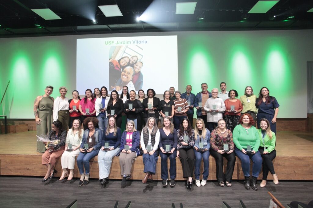 Esta é uma foto super especial de um momento de celebração! 🌟 Pelo que dá para ver, parece ser uma cerimônia de reconhecimento ou premiação, provavelmente homenageando uma equipe de saúde. Tem um grupo grande de pessoas, a maioria mulheres, reunidas no palco, algumas sentadas e outras em pé, e muitas delas estão segurando troféus ou placas de homenagem — dá para sentir o orgulho delas! O fundo tem uma iluminação verde bem vibrante e, num telão, aparece o nome "USF Jardim Vitória", o que indica que essa é a equipe que está sendo celebrada. É uma imagem que transmite muita união, felicidade e aquela sensação boa de dever cumprido. Parabéns a todos eles por esse reconhecimento!
