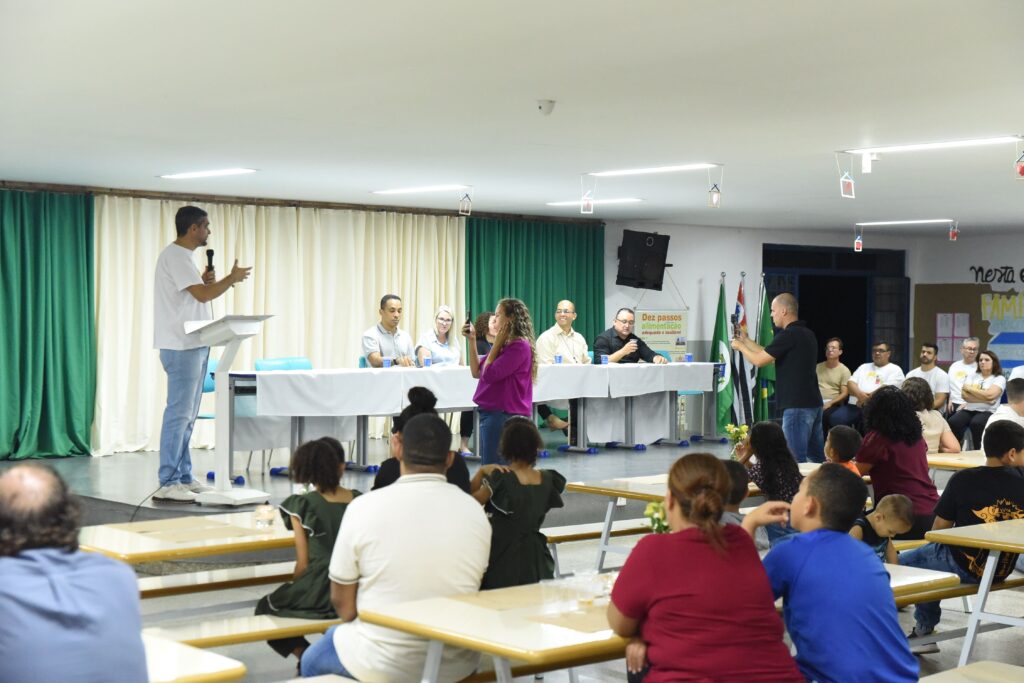 A imagem mostra um evento realizado em um ambiente interno, possivelmente uma escola ou centro comunitário, onde há uma apresentação ou palestra acontecendo. No palco, um homem está falando ao microfone, enquanto cinco pessoas estão sentadas atrás dele em uma mesa longa coberta com uma toalha branca, parecendo fazer parte da comissão organizadora ou painel de discussão. No ambiente, há várias pessoas sentadas em mesas, prestando atenção ao palestrante. Algumas crianças também estão presentes, indicando que o evento pode ser aberto a todas as idades. À direita da imagem, um homem está gravando o evento com um celular ou câmera. A decoração do local é simples, com cortinas verdes e creme ao fundo e algumas decorações penduradas no teto. Na parede ao fundo, há um cartaz com o texto "Dez passos para uma alimentação adequada e saudável", sugerindo que o tema do evento está relacionado à nutrição e saúde alimentar. A iluminação é feita por lâmpadas fluorescentes no teto, proporcionando um ambiente claro e adequado para a apresentação. A atmosfera geral é de aprendizado e compartilhamento de informações importantes para a comunidade.