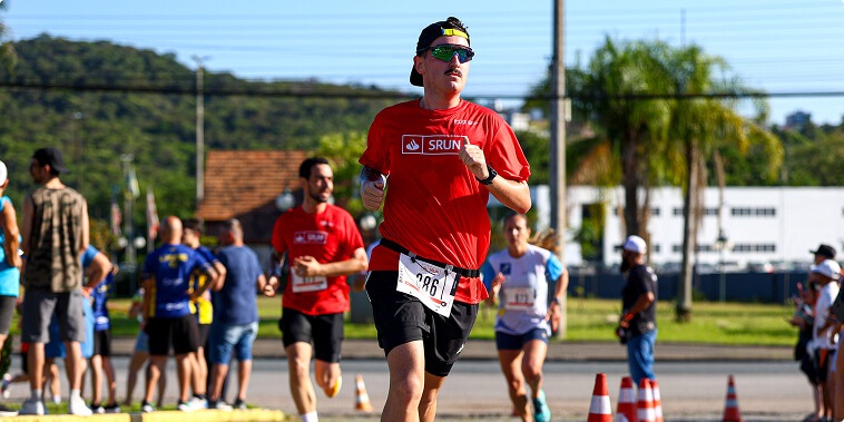 A imagem mostra um corredor masculino em destaque, participando de uma corrida ao ar livre, possivelmente uma maratona ou uma prova de 5 km. Ele veste uma camiseta vermelha com o logo "SRUN" em branco, shorts pretos, um boné preto virado para trás e óculos esportivos espelhados. O número de peito "086" está preso aos seus shorts. O corredor está em movimento, com uma expressão determinada, correndo em direção à câmera. Ao fundo, é possível ver outros corredores, incluindo um homem também com camiseta vermelha "SRUN" e uma mulher com camiseta azul clara. O cenário é ao ar livre, em um dia ensolarado, com céu azul, árvores e uma construção com telhado marrom ao fundo. Há cones laranja e branco posicionados na pista, indicando o percurso da corrida. A foto tem um foco nítido no corredor principal, enquanto o fundo está ligeiramente desfocado, destacando o sujeito principal.