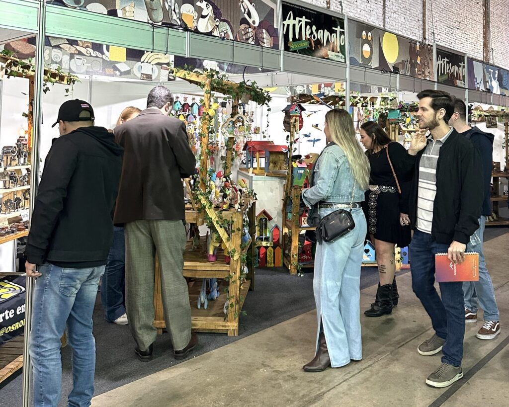 A imagem mostra uma feira de artesanato em um ambiente interno, possivelmente um galpão com paredes de tijolos aparentes. No centro da imagem, várias pessoas estão observando e interagindo com uma barraca que vende peças artesanais feitas principalmente de madeira, como casinhas para pássaros e pequenas esculturas. A barraca é decorada com trepadeiras artificiais e possui uma estrutura simples de madeira. Há um banner colorido na parte superior com a palavra "Artesanato" destacada, sugerindo que o foco do evento são produtos feitos à mão. As pessoas na imagem estão vestidas de forma casual e parecem interessadas nas peças expostas. O ambiente é bem iluminado, o que valoriza os detalhes dos objetos artesanais. A atmosfera geral transmite a sensação de um evento cultural voltado para a valorização do trabalho manual e da arte local.