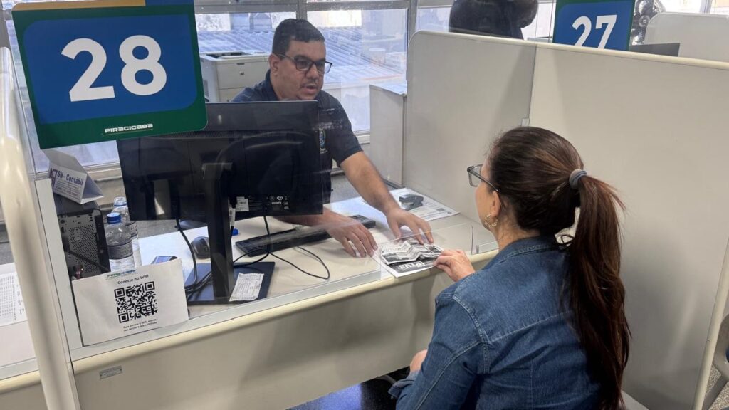 A imagem mostra um atendimento presencial em um órgão público, provavelmente a prefeitura de Piracicaba, conforme indicado pelo logotipo na placa. * À esquerda, um funcionário usa óculos e uma camisa polo escura, estando sentado em sua estação de trabalho (guichê 28). Ele interage com uma cidadã, posicionando documentos sobre o balcão. * À direita, uma mulher de costas, usando uma jaqueta jeans e prendendo o cabelo em um rabo de cavalo, está sentada à frente do funcionário, recebendo o atendimento. * Sobre a mesa, há um computador, uma garrafa de água e um aviso com um código QR para acesso ao Wi-Fi local. O ambiente é um escritório administrativo com divisórias brancas, típica de um centro de atendimento ao cidadão.