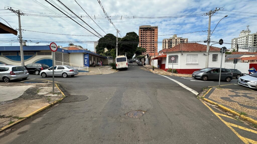 A imagem mostra uma cena urbana em um cruzamento de ruas durante o dia, com céu parcialmente nublado. À esquerda, há um prédio moderno com fachada branca e detalhes em azul, identificado como "AME" (Ambulatório Médico de Especialidades). Em frente a esse prédio, há vários carros estacionados. À direita, está uma casa residencial de estilo tradicional, com paredes brancas e faixa vermelha na parte inferior, além de telhado de telhas vermelhas. Ao fundo, é possível ver um prédio em construção, com estrutura de tijolos aparentes. A rua é asfaltada, com meio-fio pintado de amarelo e um bueiro visível no chão. Há vários fios elétricos cruzando o céu na imagem. A sinalização de trânsito inclui uma placa de "sentido único" e uma placa de "proibido entrar". Carros estão circulando ou estacionados nas vias. O ambiente transmite a sensação de um bairro misto entre residencial e comercial, com obras em andamento e infraestrutura urbana típica.