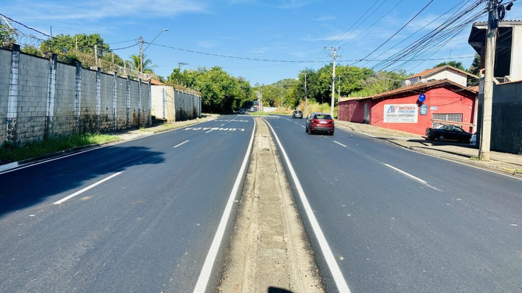 A imagem mostra uma rua asfaltada vista do ponto de vista de quem está dentro de um veículo, olhando para frente. A via possui uma faixa divisória central pintada de branco. À esquerda, há um muro alto de blocos de concreto com arame farpado no topo, indicando uma área protegida ou privada. O muro apresenta manchas escuras na base e vegetação rasteira verde ao longo dele. À direita, há um prédio vermelho com uma placa que diz "ANTENAS PIRACICABA SERVIÇOS PROFISSIONAIS" junto com dois números de telefone. Na frente do prédio, alguns carros estão estacionados, e mais à frente, na rua, há outros veículos em movimento. O céu está claro, azul e com poucas nuvens, indicando um dia ensolarado. Linhas de energia elétrica cruzam o céu acima da rua e dos edifícios. A rua segue em linha reta e depois se eleva um pouco, desaparecendo entre árvores ao fundo. A iluminação é forte, típica de meio-dia, com sombras nítidas. A cena transmite uma sensação de área urbana ou suburbana, limpa e organizada.