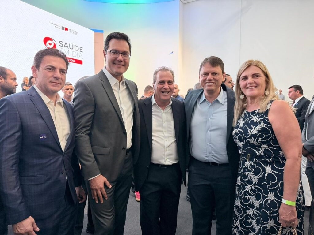 A imagem mostra um grupo de cinco pessoas posando para uma foto em um evento formal, provavelmente relacionado à saúde pública, dado o logotipo e o texto "SAÚDE EM DIA" e "SÃO PAULO" no fundo. As pessoas estão alinhadas, olhando para a câmera, com expressões sérias ou discretamente sorridentes. Quatro homens estão vestidos com ternos escuros, e uma mulher está usando um vestido azul com estampa branca. O ambiente parece ser interno, com iluminação clara e uniforme. Ao fundo, há outras pessoas desfocadas, indicando que o local está movimentado. A imagem transmite uma atmosfera profissional e oficial, possivelmente durante uma cerimônia ou anúncio importante na área da saúde.
