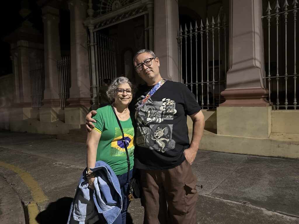 A imagem mostra um casal adulto posando para uma foto à noite em frente a um prédio clássico com uma grade de ferro ornamentada. A mulher está à esquerda, tem cabelo curto e grisalho, usa óculos, uma camiseta verde com a bandeira do Brasil estampada e calça jeans azul, segurando uma jaqueta jeans na mão esquerda. O homem está à direita, também usa óculos, veste uma camiseta preta com estampa dos personagens Tom e Jerry, calças cargo marrons e uma alça colorida atravessada no corpo, possivelmente de uma bolsa ou câmera. Eles estão próximos um do outro, com o braço do homem apoiado nos ombros da mulher. O fundo mostra uma construção clara, com colunas e uma grade alta de ferro com pontas afiadas, iluminada por uma luz artificial contra o céu escuro da noite. A iluminação da foto parece ser de flash, criando sombras definidas atrás do casal.