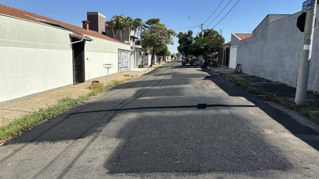 O Trabalho: É possível ver uma área recém-asfaltada com cor preta e textura rugosa, contrastando com o asfalto antigo. Equipamento: Um pequeno rolo compactador amarelo está estacionado ao fundo, indicando que a obra está em andamento ou foi concluída recentemente. Local: Trata-se de uma rua de bairro com casas, muros brancos e cinzas, e uma placa de sinalização indicando o nome "ATHAULPA VAZ DE MELLO" em um poste. Clima: O dia está limpo, com céu azul e uma luz solar forte que projeta sombras bem definidas.