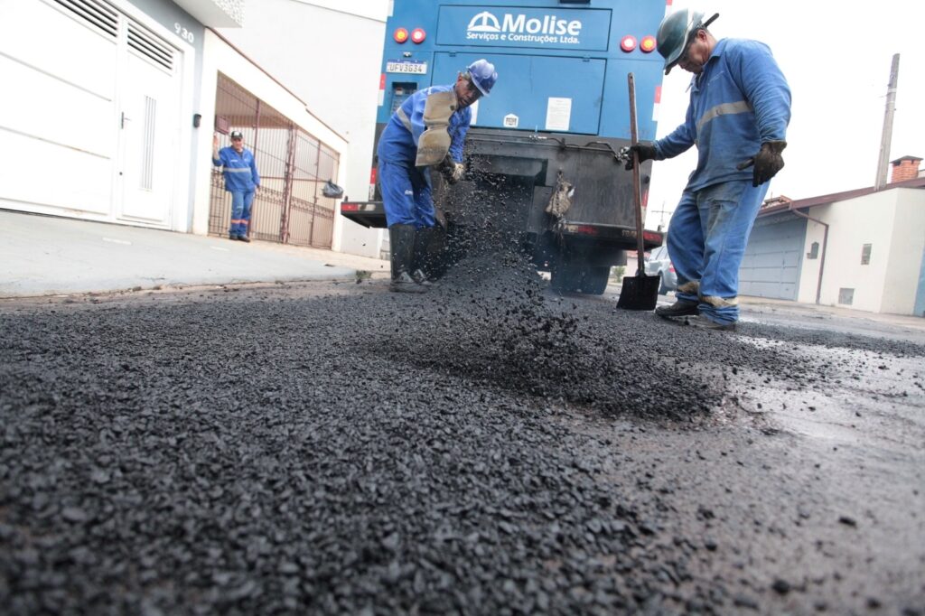 A imagem mostra uma equipe de trabalhadores realizando uma obra de pavimentação asfáltica em uma rua residencial. * Em primeiro plano, vê-se uma grande quantidade de massa asfáltica fresca e granulada sendo espalhada sobre a via. * Dois operários, vestidos com uniformes azuis de proteção, capacetes e botas, estão em plena atividade: um deles manuseia uma pá para distribuir o material, enquanto o outro auxilia no processo próximo à parte traseira de um caminhão. * O caminhão, que pertence à empresa "Molise Serviços e Construções Ltda.", está estacionado na via e serve de suporte para o trabalho. * Ao fundo, à esquerda, um terceiro trabalhador observa a operação, enquanto casas e portões residenciais compõem o cenário urbano ao redor.