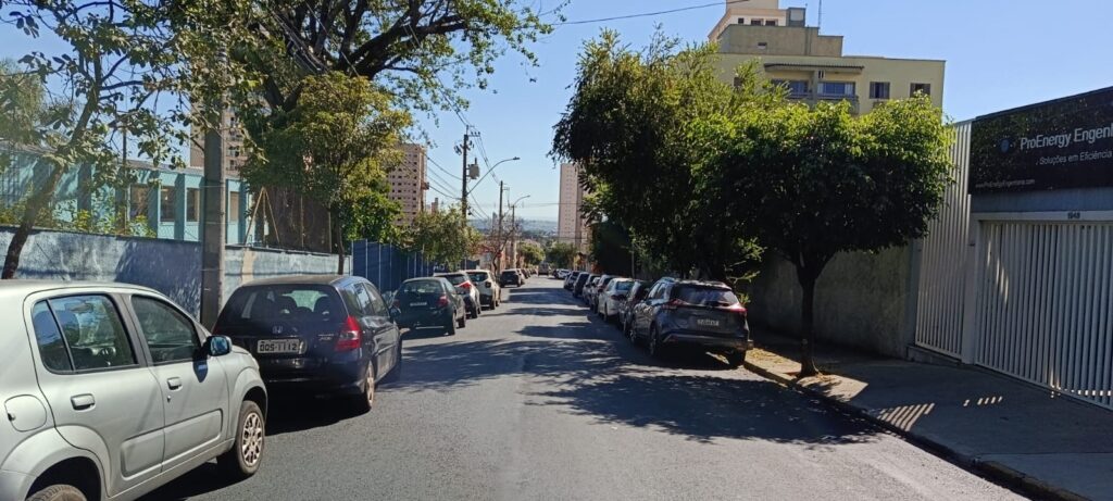 A imagem mostra uma rua urbana em um dia ensolarado, com uma perspectiva em direção ao fundo. * O lado esquerdo da rua apresenta um muro azul, árvores altas e dois veículos estacionados em primeiro plano: um carro prateado e um Honda Fit escuro. * O lado direito é composto por uma calçada com árvores e o muro de uma empresa chamada "ProEnergy Engenharia", com seu letreiro visível. * Há uma fileira de carros estacionados ao longo de ambos os lados da via, que segue em declive. * Ao fundo, é possível observar outros prédios e árvores, sugerindo uma área residencial ou comercial de densidade média. O céu está limpo e azul, indicando condições climáticas claras.
