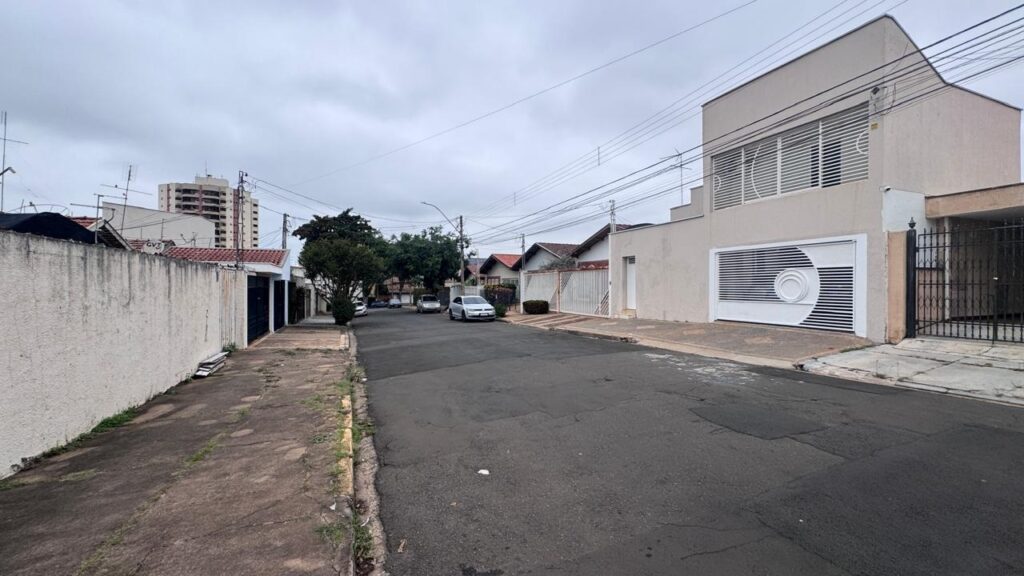 A imagem mostra uma rua residencial tranquila em um dia nublado. A rua é pavimentada e está ladeada por calçadas. Do lado direito, há uma casa de dois andares com fachada clara, portão branco com detalhes horizontais e um elemento circular decorativo. Do lado esquerdo, vê-se um muro de concreto simples que acompanha a calçada. Ao longo da rua, alguns carros estão estacionados. O céu está coberto por nuvens, o que deixa a iluminação mais suave e difusa. Há fios de eletricidade cruzando o céu, e a atmosfera geral é calma e silenciosa, típica de um bairro residencial.