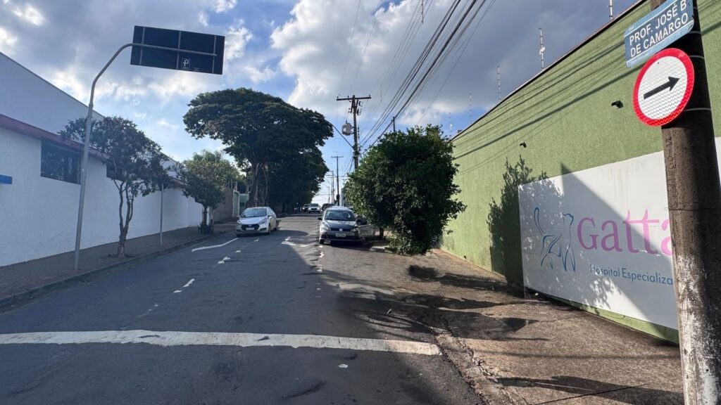 A imagem mostra uma rua urbana durante o dia, vista a partir do nível da rua, com uma perspectiva que leva o olhar para frente, ao longo da via. À direita, há um muro verde com uma placa que indica "Gatta Hospital Especializado", acompanhada de um desenho de um gato. Também à direita, há um poste de luz com uma placa de rua que diz "RUA PROF. JOSÉ B. DE CAMARGO" e uma placa de trânsito com uma seta indicando para a direita. Dois carros estão circulando pela rua, que é asfaltada e tem faixas de pedestres visíveis. Árvores ao longo da via proporcionam sombra, e o céu está limpo, com algumas nuvens brancas, sugerindo um dia ensolarado. A cena é tranquila, típica de uma área urbana com comércio e serviços.