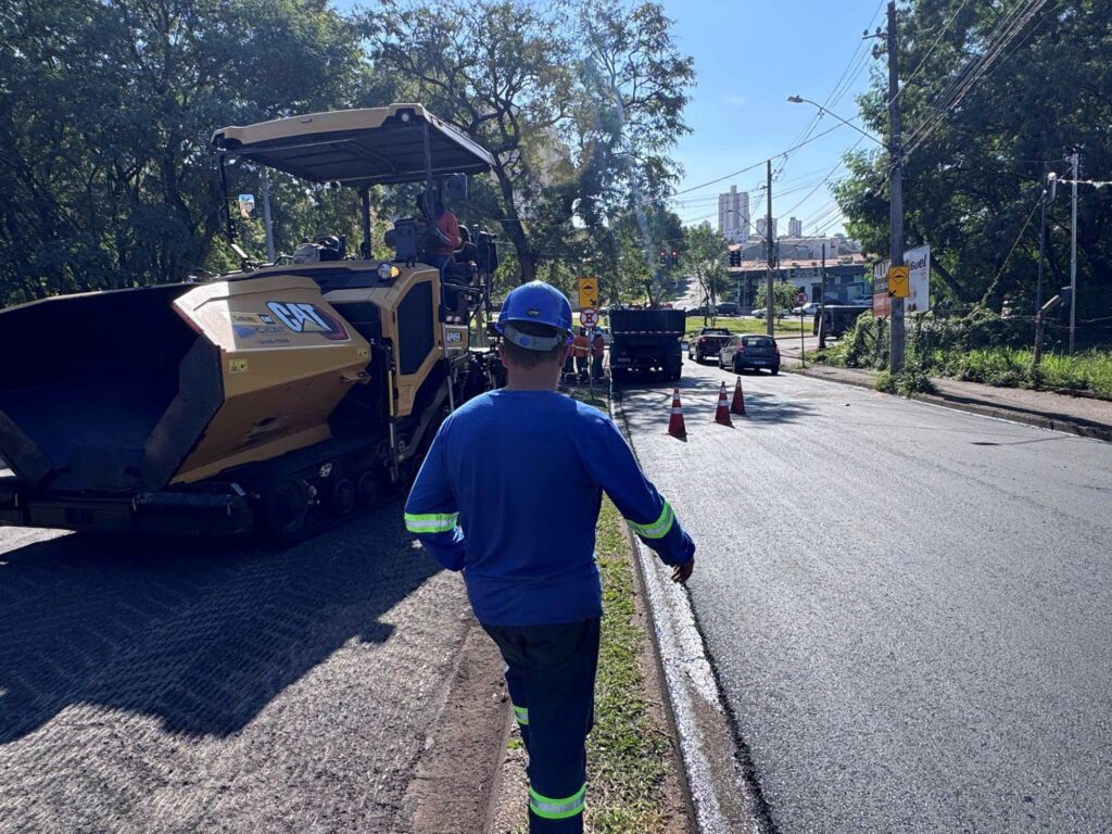 . Ela mostra uma cena bem comum de manutenção urbana em um dia ensolarado. O destaque principal é um operário que está de costas, caminhando perto da via; ele está equipado com um uniforme azul cheio de faixas refletivas e um capacete de segurança azul. Bem ali do lado dele, tem uma daquelas máquinas pavimentadoras da CAT, bem grandes e amarelas, que estão fazendo o serviço de recapeamento. A rua tem um asfalto bem escuro e novo, e a área está sinalizada com vários cones laranjas. O lugar parece ser bem arborizado, com árvores frondosas dos dois lados, e lá no fundo, dá para ver um pouquinho do horizonte urbano com alguns prédios. Parece ser uma operação de rotina para deixar a rua novinha em folha!