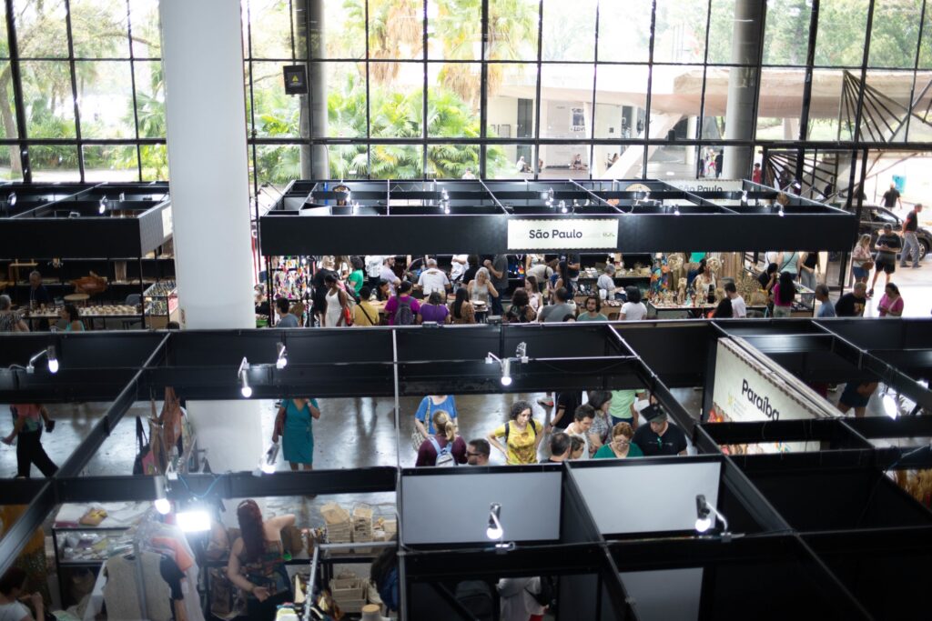 A imagem mostra uma feira ou mercado interno bastante movimentado, visto de cima. O espaço é organizado em várias barracas estruturadas com armações metálicas pretas, formando uma espécie de grade que divide o ambiente em corredores. Muitas pessoas circulam pelos corredores, observando e interagindo com os produtos expostos. O local parece ser um grande salão com teto alto e janelas amplas, por onde entra bastante luz natural. Do lado de fora, é possível ver árvores e plantas, o que traz um contraste agradável entre o ambiente interno e a natureza externa. As barracas exibem uma variedade grande de artigos, como artesanato, roupas, bolsas, bijuterias e objetos decorativos. Algumas placas indicam nomes de regiões brasileiras, como "São Paulo" e "Paraíba", sugerindo que a feira reúne produtos típicos dessas localidades. O ambiente transmite uma sensação de diversidade cultural e comercial, com muitas pessoas interessadas em conhecer e comprar os itens oferecidos. De modo geral, a imagem retrata um evento vibrante, cheio de movimento e cores, que celebra a cultura e o comércio regional em um espaço amplo e bem iluminado.