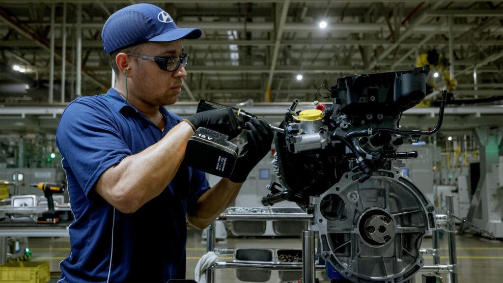 A imagem mostra um operário em uma linha de montagem da Hyundai, trabalhando em um motor de carro. Ele está usando um boné azul com o logo da Hyundai, óculos de proteção, luvas pretas e uma camisa polo azul. O homem está utilizando uma ferramenta elétrica de 18 volts para montar ou apertar componentes do motor, que está posicionado em um suporte metálico à sua frente. O ambiente é uma fábrica industrial, com iluminação forte e estrutura metálica visível no teto. A cena transmite uma sensação de concentração, profissionalismo e precisão no trabalho.