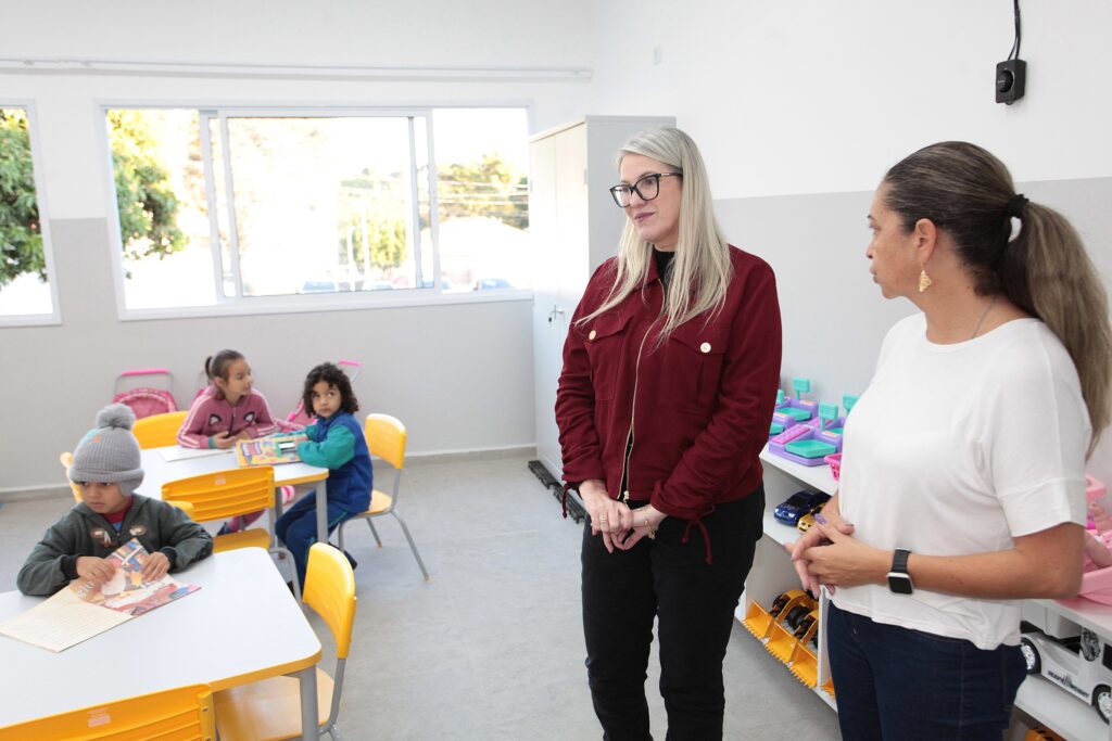 A imagem mostra uma sala de aula de educação infantil iluminada e organizada, com duas mulheres adultas em pé, conversando no lado direito. * A mulher à esquerda veste uma jaqueta vermelha, calça preta e óculos. A mulher à direita usa uma blusa branca, calça jeans e um relógio inteligente no pulso. * No lado esquerdo, três crianças estão sentadas em mesas amarelas, concentradas em atividades. Uma criança usa um gorro cinza e lê um livro, enquanto as outras duas, uma menina de blusa rosa e outra de blusa azul, estão sentadas à mesa ao fundo. * O ambiente possui janelas amplas que permitem a entrada de luz natural, piso claro e prateleiras organizadas com brinquedos e materiais pedagógicos, sugerindo um espaço de aprendizado bem equipado e acolhedor.