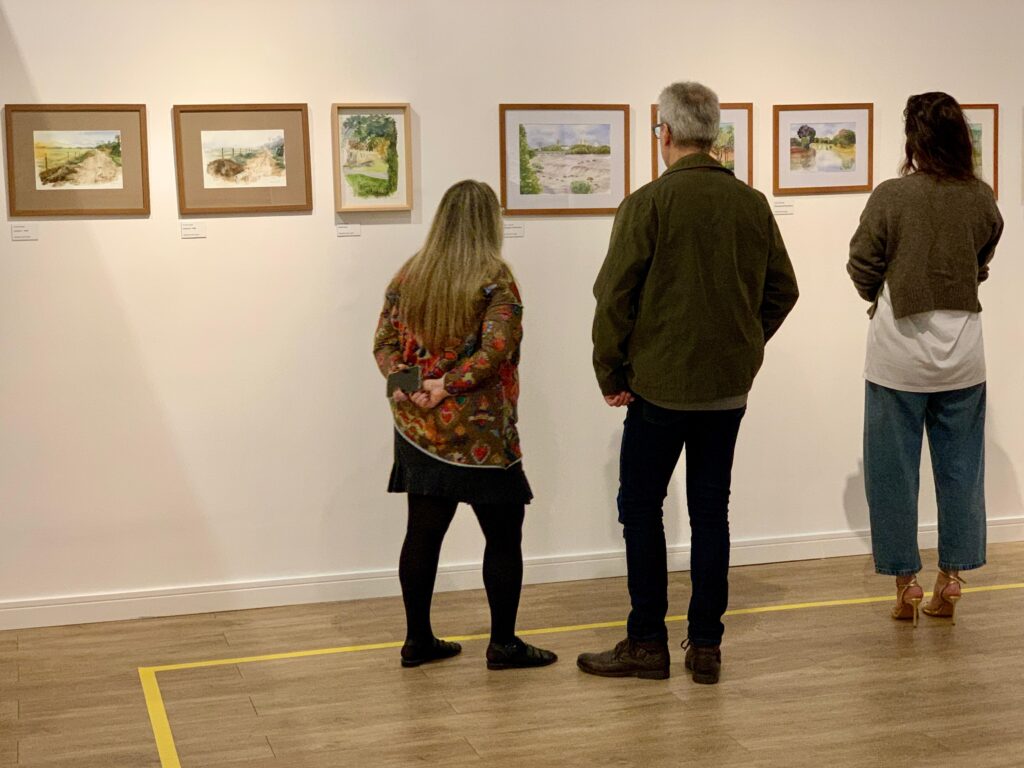 A imagem mostra três pessoas de costas, observando uma exposição de arte em uma galeria. * A pessoa à esquerda veste uma blusa estampada colorida, saia preta e meia-calça. * A pessoa no centro usa uma jaqueta verde-oliva, calça jeans escura e sapatos marrons. * A pessoa à direita veste um suéter marrom, camiseta branca, calça jeans larga e sandálias de salto alto. Eles estão em frente a uma parede branca onde estão pendurados vários quadros com pinturas em aquarela emolduradas em madeira. O chão é de madeira clara e há uma fita amarela demarcando uma área de segurança no piso.