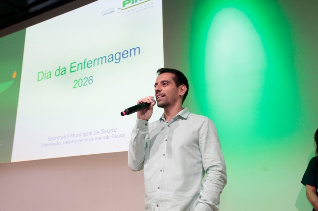 A imagem mostra um homem falando em um microfone, provavelmente durante um evento ou palestra. Ele está vestido com uma camisa azul clara e parece estar em um palco ou em um ambiente de conferência. Ao fundo, há uma tela grande com a inscrição "Dia da Enfermagem 2026", além dos nomes "Secretaria Municipal de Saúde" e "Organização: Departamento de Atenção Básica". A parede atrás da tela é verde, iluminada por uma luz que cria um efeito de destaque. O homem está sorrindo levemente e olhando para o lado, sugerindo que está se comunicando com o público. A atmosfera parece formal e profissional, relacionada a uma celebração ou evento oficial da área da saúde.