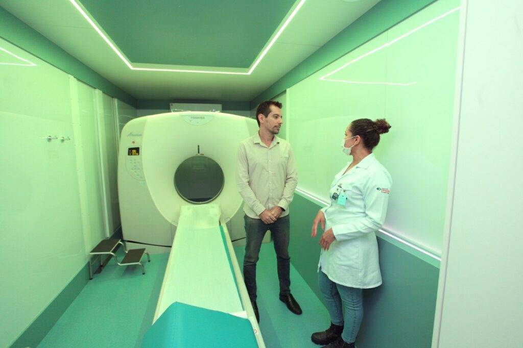 A imagem mostra um ambiente clínico moderno, provavelmente uma sala de tomografia computadorizada (TC). No centro da imagem, há um homem vestido com camisa clara e jeans, conversando com uma profissional da saúde, que usa jaleco branco, máscara facial e botas escuras. Eles estão próximos a um equipamento de tomografia da marca Toshiba, modelo Alexion, que é grande, branco e possui uma abertura circular onde o paciente é colocado para o exame. A sala é bem iluminada, com paredes em tons claros e detalhes em verde menta, transmitindo um ambiente limpo e organizado. Também é possível ver um pequeno banquinho metálico próximo ao equipamento. A cena sugere uma interação profissional, possivelmente explicando o procedimento do exame.