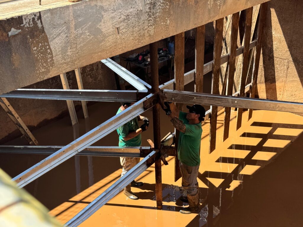 A imagem mostra dois trabalhadores dentro de uma estrutura de concreto, provavelmente um canal ou sistema de drenagem, com água barrenta até a altura dos joelhos. Eles estão usando botas de borracha e camisetas verdes. Um dos homens, mais próximo da câmera, está segurando e fixando uma viga metálica a um poste vertical central, enquanto o outro, ao fundo, também está envolvido na montagem ou manutenção da estrutura metálica. A cena é iluminada pela luz do sol, que cria sombras marcantes das vigas no interior do canal. Acima deles, há uma ponte ou passarela de concreto. A imagem transmite uma sensação de trabalho manual intenso em um ambiente industrial e úmido.