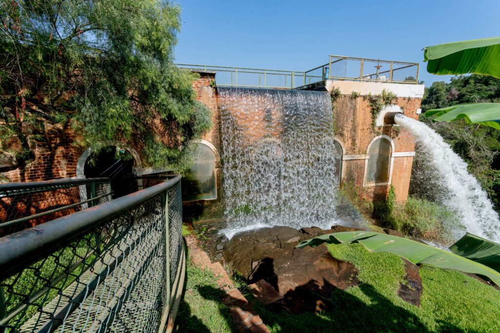 Esta imagem mostra uma estrutura arquitetônica histórica de tijolos aparentes, que integra uma queda d'água artificial em sua fachada. * O edifício possui arcos que lembram janelas ou passagens e é coroado por uma grade de proteção metálica no topo. * À direita, um grande cano projeta um forte jato de água, que se une ao fluxo que cai verticalmente pela parede da construção. * Em primeiro plano, à esquerda, há uma passarela com uma grade de proteção de tela metálica. * O cenário é cercado por vegetação, incluindo árvores e folhas de bananeira que emolduram a cena, criando um contraste entre a engenharia humana e a natureza.