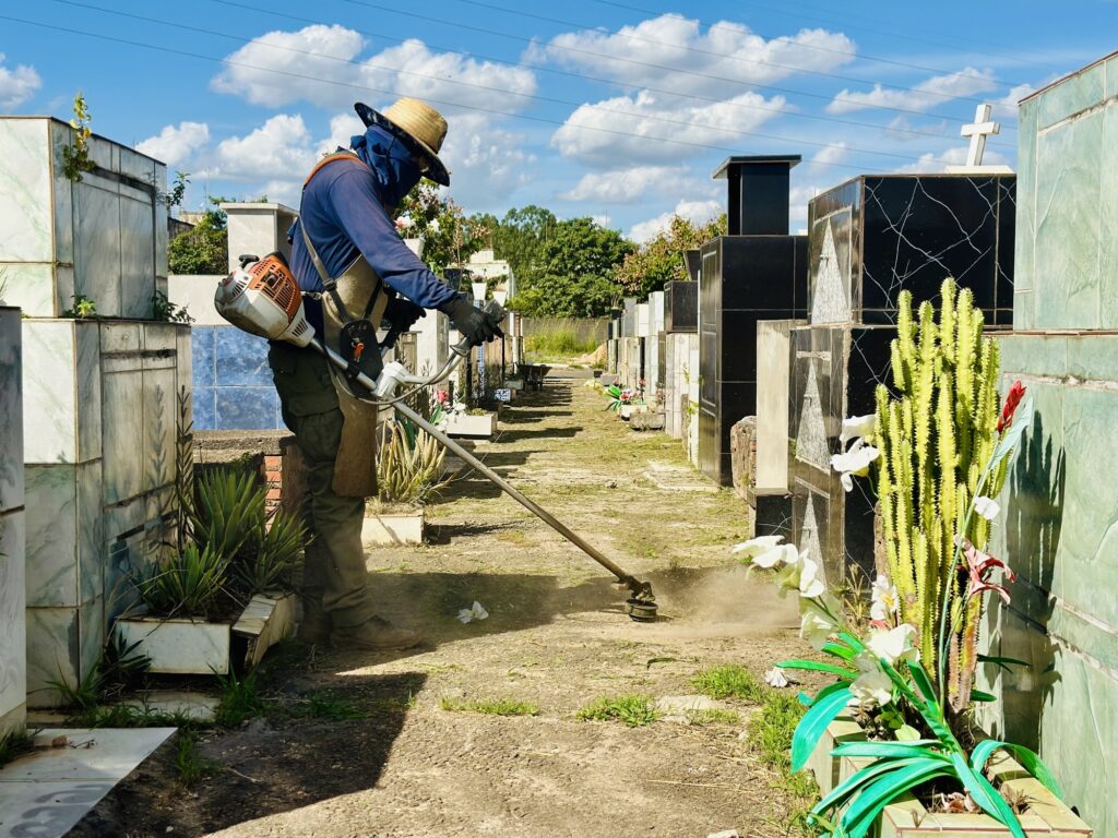 A imagem mostra um zelador cuidando da manutenção de um cemitério em um dia ensolarado. Ele está usando uma camisa azul de manga longa, calças verdes, um chapéu de palha e uma máscara facial azul, enquanto utiliza um aparador de grama para cortar a vegetação que cresce entre os túmulos. O cemitério é composto por várias construções de mármore, algumas com cruzes e flores decorativas, organizadas em fileiras ao longo de um caminho de terra. A vegetação ao redor é verde e abundante, e há vasos com plantas, incluindo um cacto com flores artificiais brancas e vermelhas. O céu está parcialmente nublado, e a luz do sol forte cria sombras definidas no solo e destaca a poeira levantada pela ferramenta de corte. O ambiente transmite uma sensação de cuidado e respeito, mostrando o trabalho dedicado para manter o local limpo e organizado. A imagem tem uma boa qualidade, com foco claro no zelador e na ação que ele realiza.