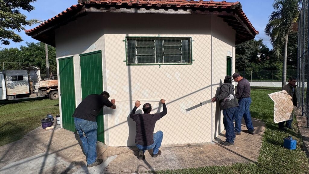 A imagem mostra quatro homens trabalhando na parte externa de um pequeno edifício hexagonal feito de alvenaria. O prédio é pintado de bege claro e possui portas e uma janela com grade de segurança na cor verde. Dois homens estão próximos ao centro da parede, aparentemente medindo ou marcando algo; um está em pé e o outro agachado. À direita, outros dois homens estão usando uma régua de nível longa contra a parede, provavelmente para garantir que a superfície esteja nivelada. O chão é de concreto e há áreas de grama ao redor. Ao fundo, é possível ver um caminhão branco estacionado e uma cerca de arame, indicando que o local pode ser um parque ou uma área de lazer. A cena ocorre durante o dia, com luz solar forte que projeta sombras da cerca na parede do edifício. A foto foi tirada em ângulo amplo, capturando claramente o ambiente e o trabalho em andamento.