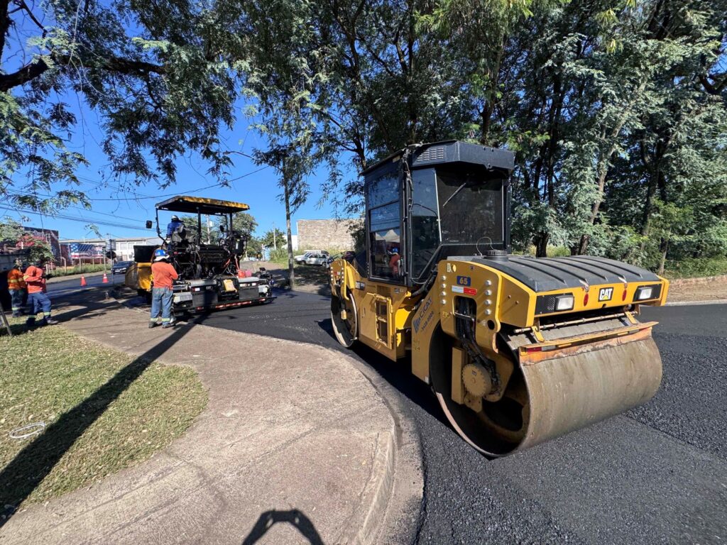 A imagem mostra uma cena de construção ou manutenção de estrada em um dia ensolarado. Em primeiro plano, há um rolo compactador amarelo da marca Caterpillar (CAT) estacionado sobre uma camada de asfalto recém-aplicada. À esquerda, um pouco mais ao fundo, está uma pavimentadora de asfalto também amarela da Caterpillar, com um trabalhador vestido com camiseta laranja e capacete azul próximo a ela. Outro trabalhador, com vestimenta semelhante, aparece mais ao fundo à esquerda. A estrada está ladeada por uma calçada de concreto e uma faixa de grama verde, enquanto árvores altas e frondosas proporcionam sombra ao fundo. A iluminação é natural e intensa, típica de um dia claro. A imagem transmite a ideia de um trabalho de infraestrutura em andamento, com equipamentos pesados e trabalhadores especializados.