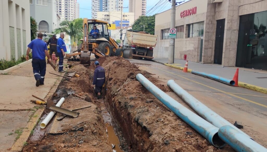 Esta imagem mostra uma obra de infraestrutura urbana em andamento. O destaque principal é uma vala aberta na rua, com uma retroescavadeira amarela e uma equipe trabalhando na instalação de tubulações de saneamento ou drenagem. A área está sinalizada com cones de segurança e é possível observar tubos de PVC azuis aguardando instalação, além de um caminhão caçamba para o transporte de terra. É uma cena típica de reparos em redes de água ou esgoto em uma via pública.